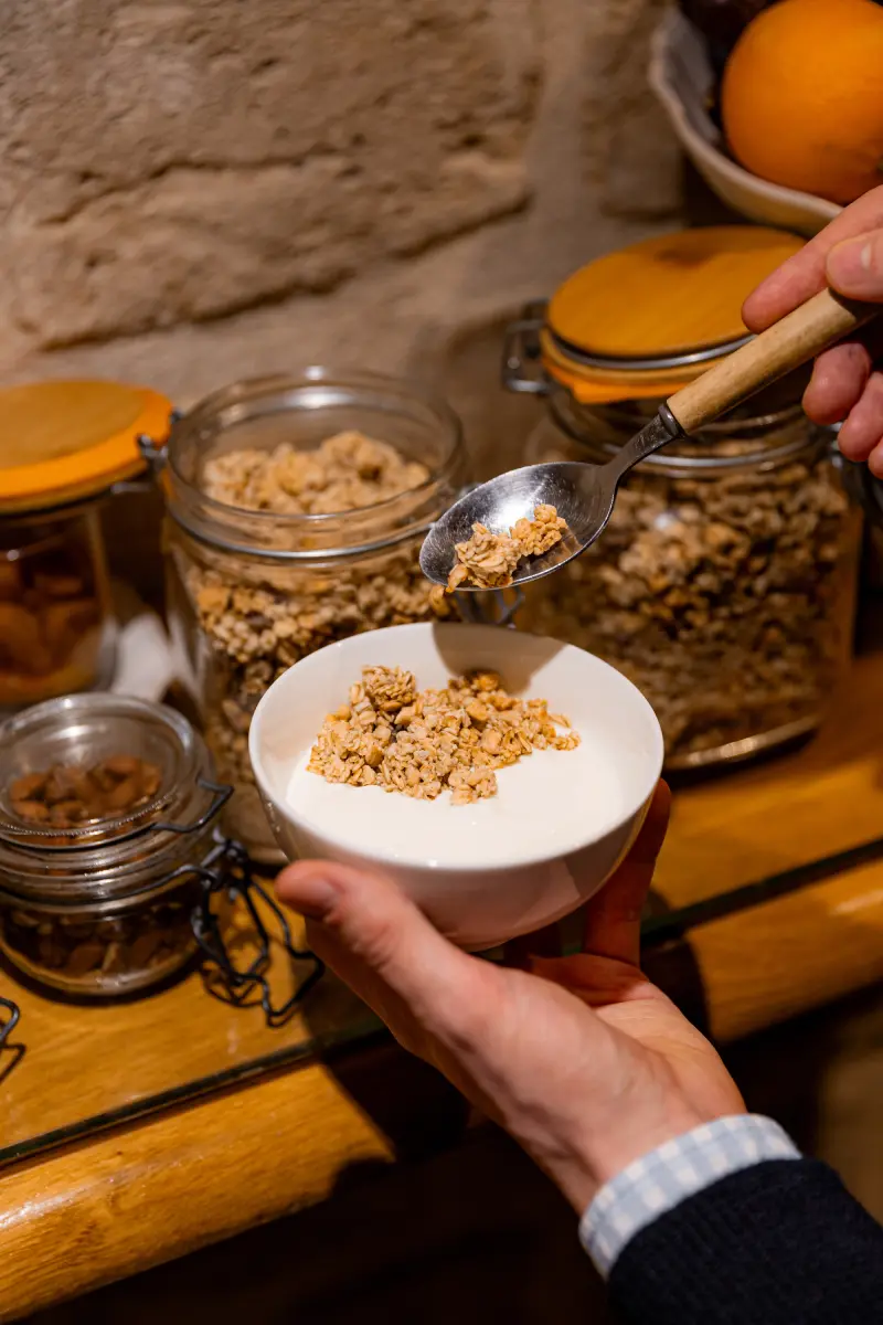 Mains tenant un bol de yaourt garni de granola verse a la cuillere, bocaux de cereales et oranges en arriere-plan