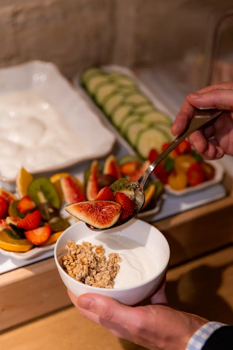 Mains preparant un bol de granola et yaourt avec plateau de fruits frais (figues, kiwi, fraises) en arriere-plan